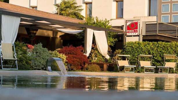Área de piscina de hotel com espreguiçadeiras, cascata e placa com 'POSTA 77'.