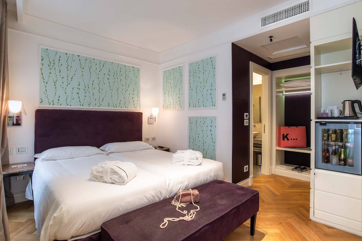 A neatly made bed with white linens, a purple bench with a pearl necklace, and a minibar with a red bag.