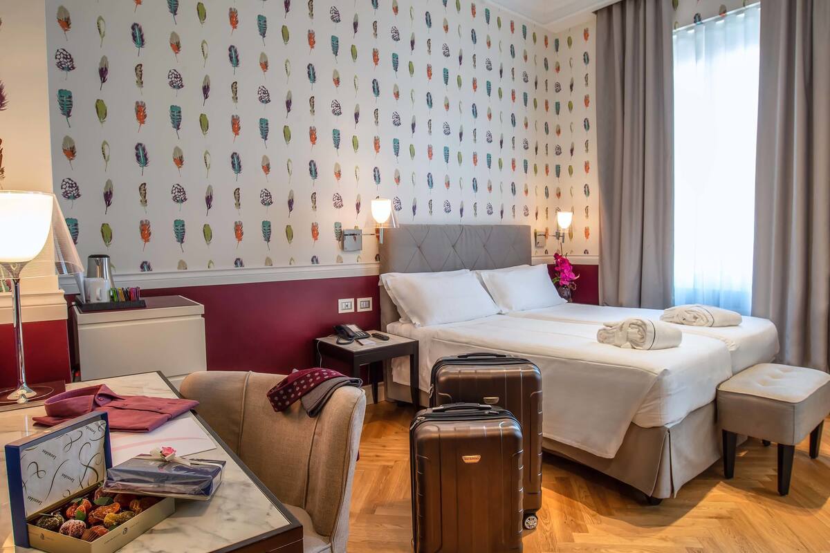 A hotel room with a large bed, a desk with a lamp, a chair, a small table with a box of chocolates, and a suitcase.