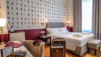 Una camera d'albergo con un letto grande, una scrivania con una lampada, una sedia, un tavolino con una scatola di cioccolatini e una valigia.