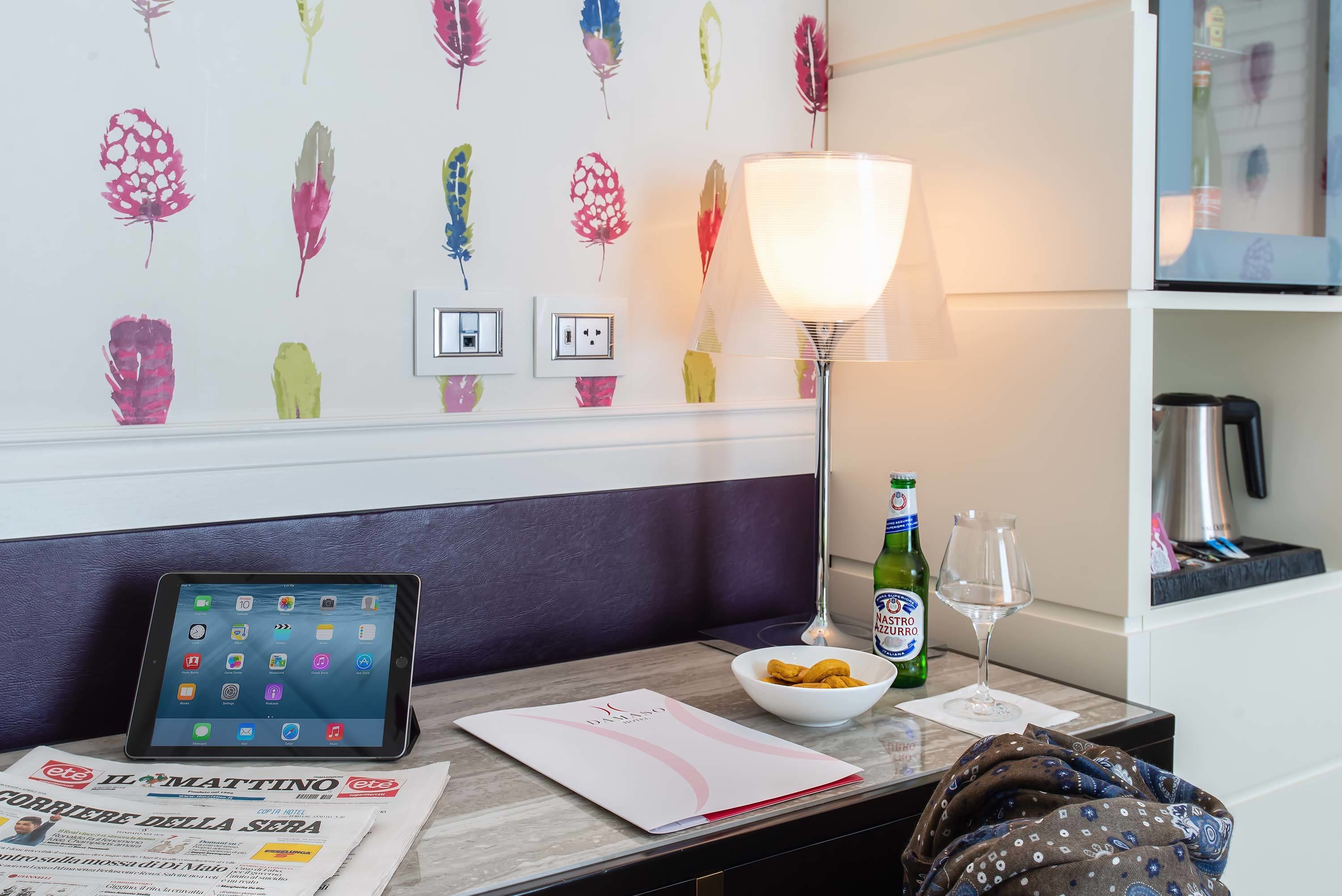 A hotel room with a tablet on a stand, a bowl of snacks, a wine glass, and a lamp on the nightstand.