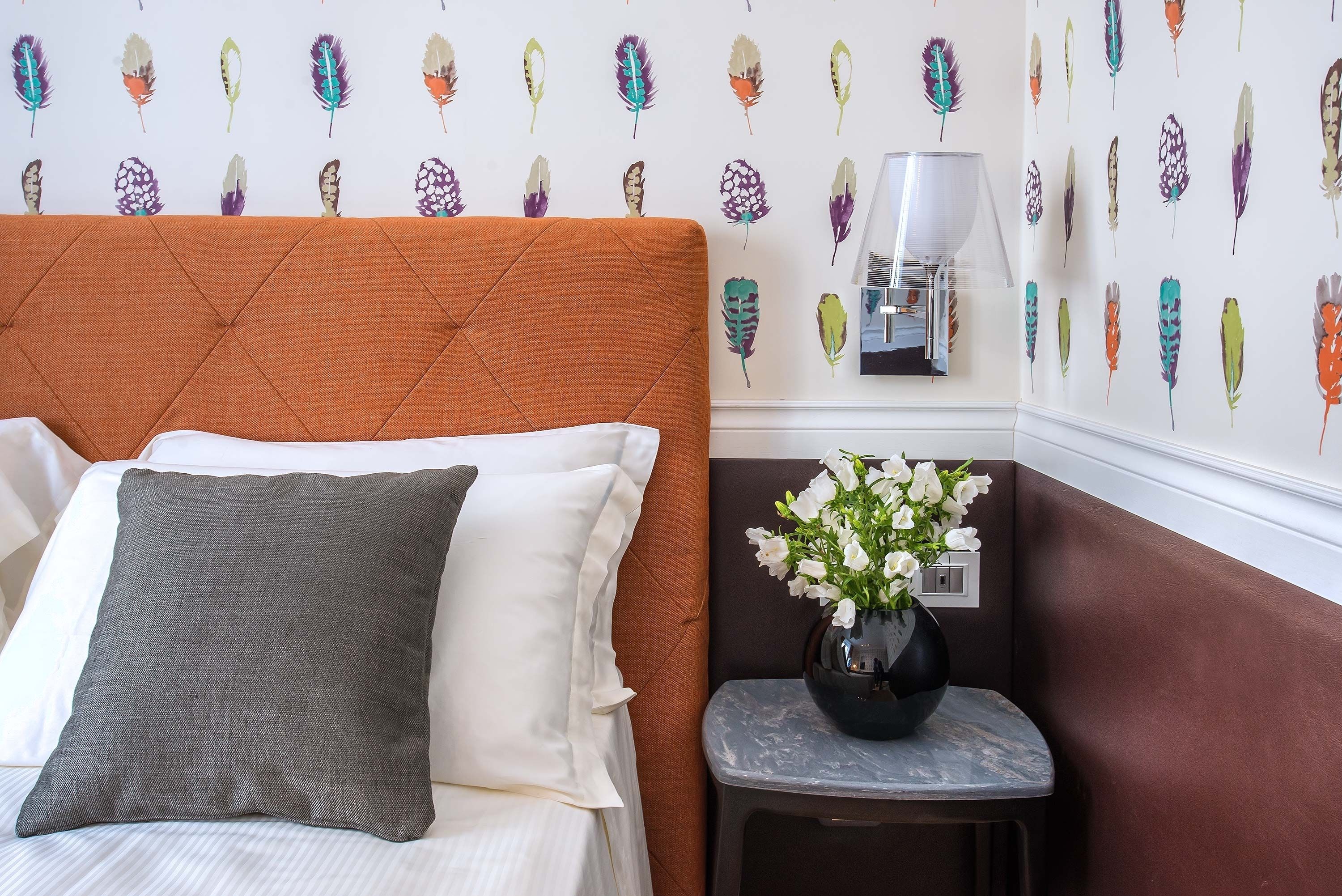Una cama bien hecha con un almohadón gris y uno blanco, un cabecero tapizado en marrón, una mesita con un florero de flores blancas, y una pared con diseño de plumas.