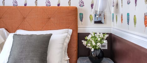 Una cama bien hecha con un almohadón gris y uno blanco, un cabecero tapizado en marrón, una mesita con un florero de flores blancas, y una pared con diseño de plumas.