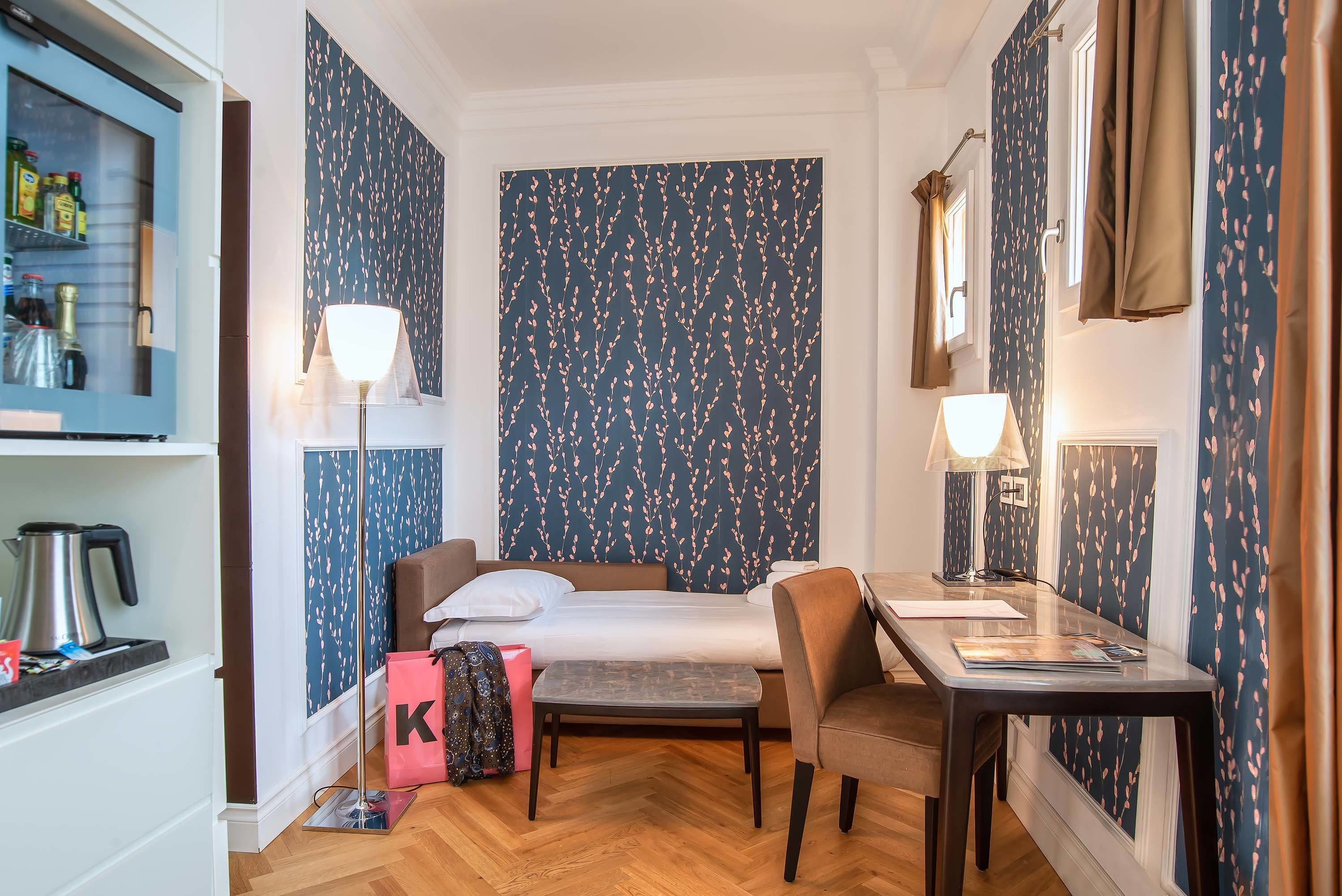 Ett hotellrum med en säng, ett skrivbord, en stol, en lampa, en vattenkokare och en resväska.