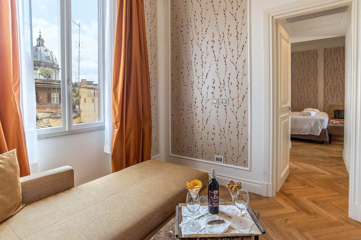 A hotel room with a sofa, a tray with wine, glasses, and snacks, a view of a cityscape through a window, and a door leading to another room.