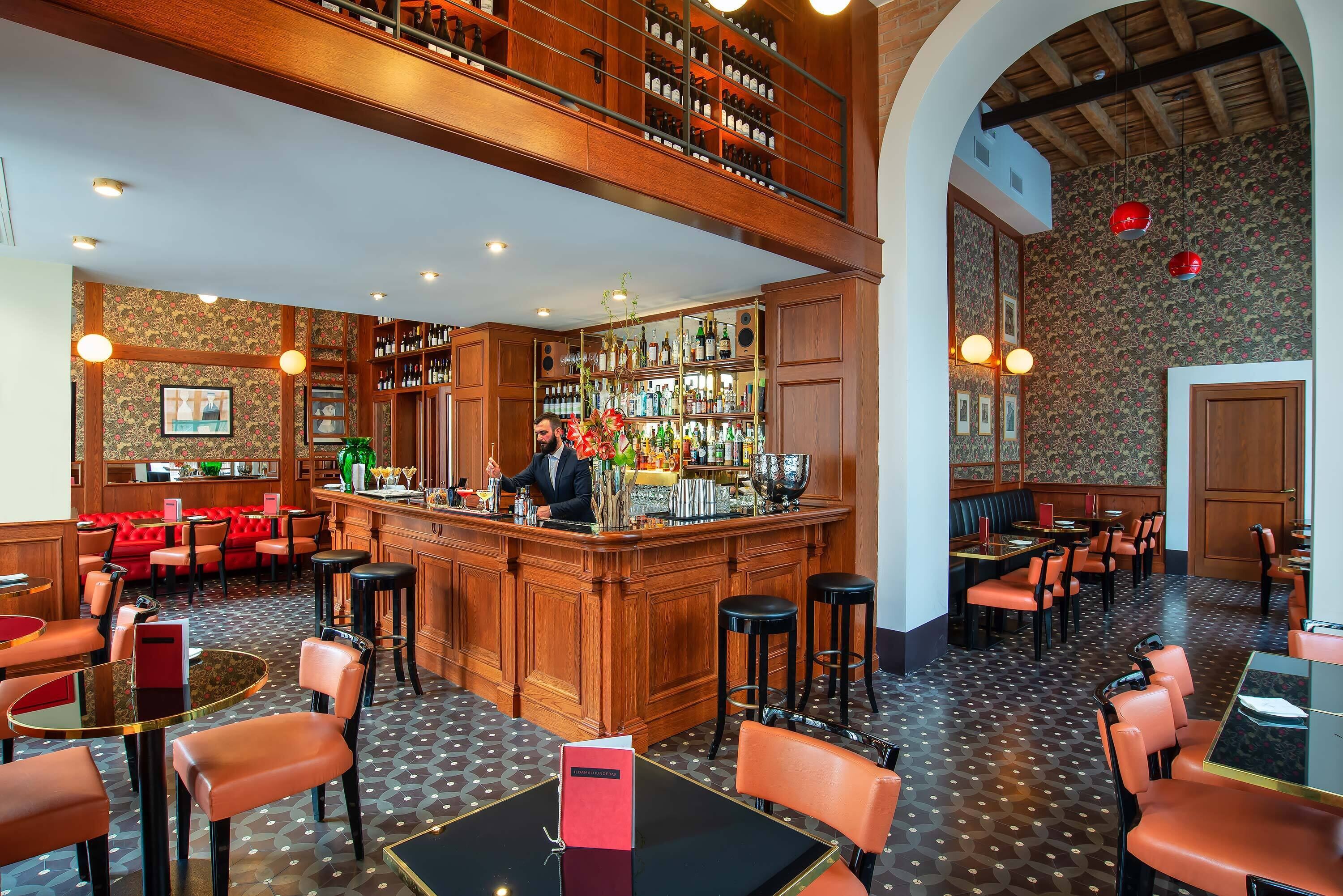 Zona de bar con un cantinero, mostrador de madera y estantes llenos de botellas. Hay cabinas tapizadas en rojo y mesas con sillas.
