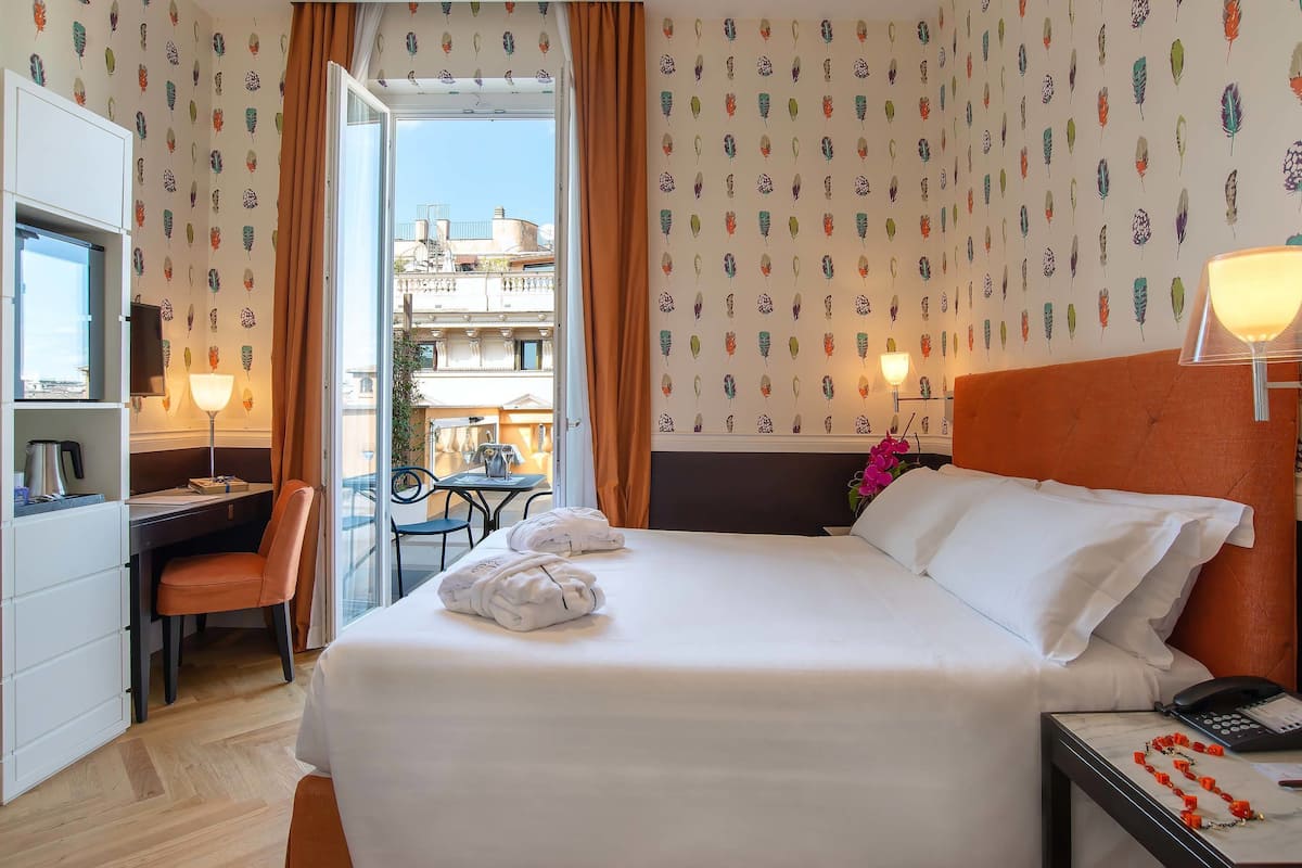 A hotel room with a bed, desk, chair, and a view of the city through the window.