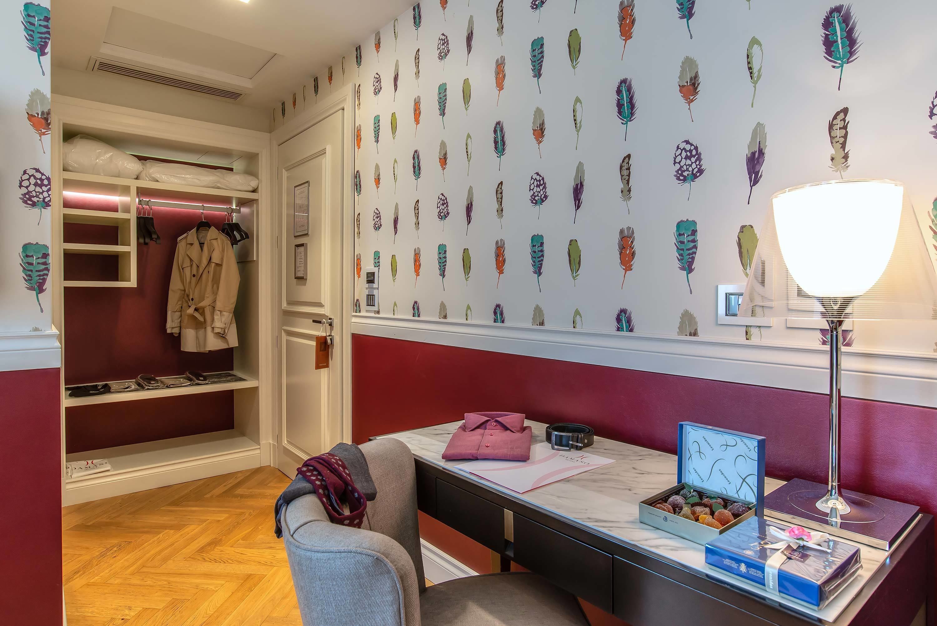 Une chambre d’hôtel avec un placard, un bureau avec une lampe, une chaise, une table avec une boîte de chocolats et un mur orné d’un motif de plumes.
