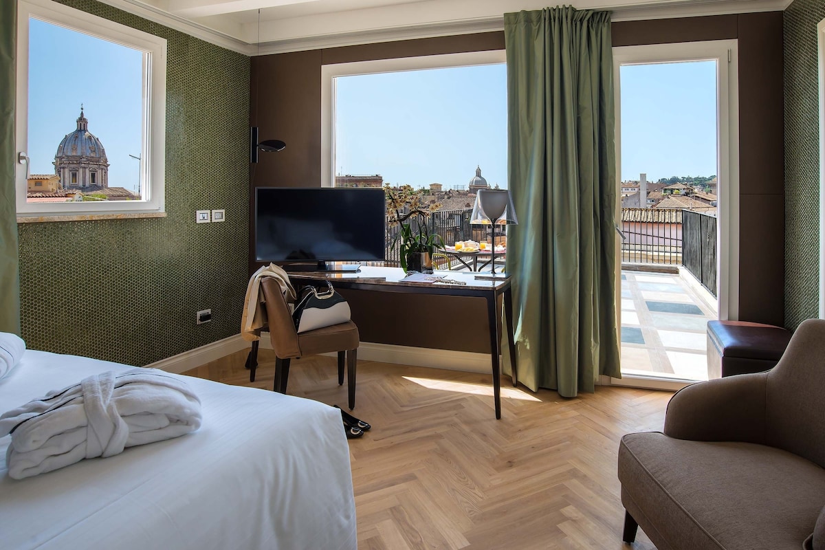 A hotel room with a bed, a desk with a TV, a chair, and a balcony with a view of the city.