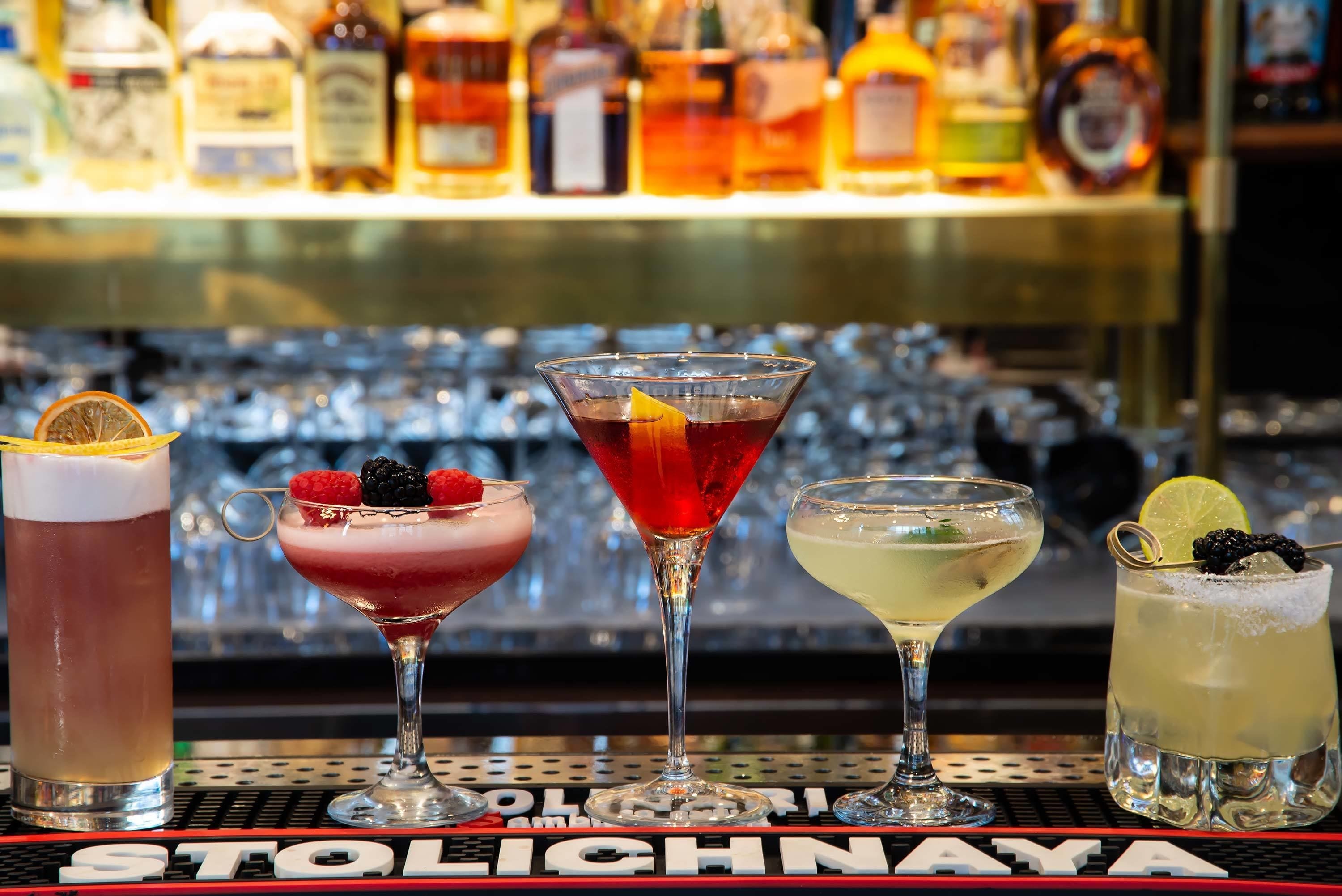 A bar with four different cocktails, each garnished with fruits and a slice of lime. Behind the bar, there are shelves with various bottles of alcohol.