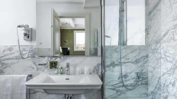 A bathroom with a marble sink, a handheld hair dryer, a glass-enclosed shower, and a mirror reflecting a green-tiled room.