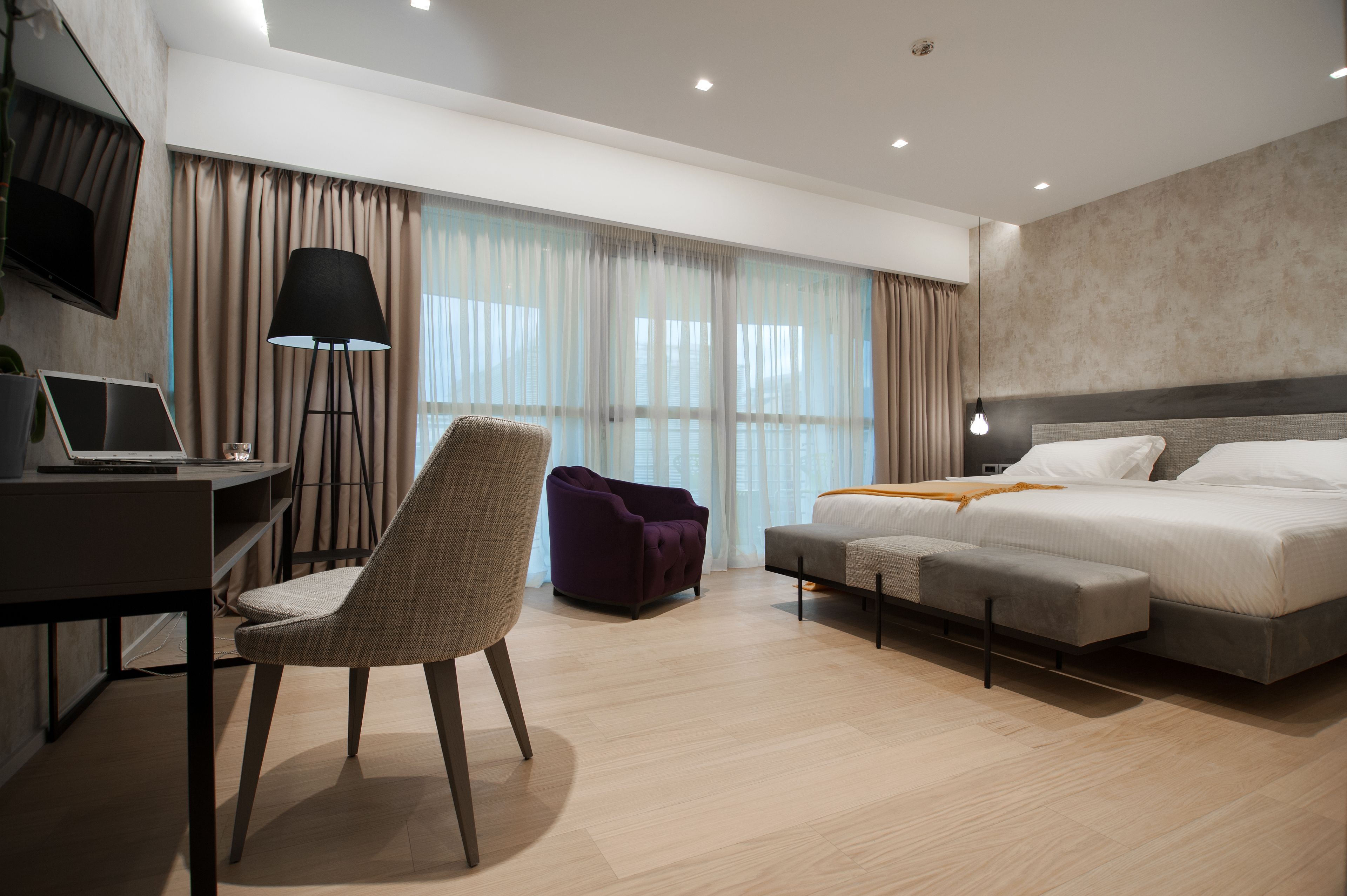 A modern hotel room with a bed, desk, chair, and large windows.