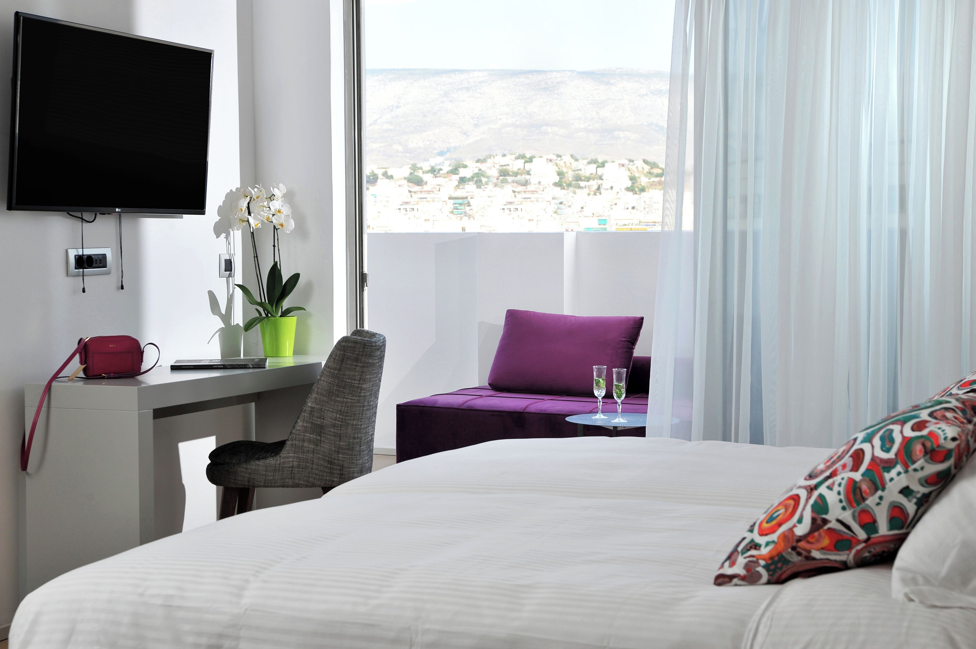 Habitación de hotel con cama, escritorio con televisor, silla, almohada morada y vista a un paisaje urbano.