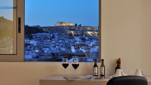 A room with a large window overlooking a cityscape, two wine glasses, and a bottle of wine on a table. - Athenaeum Grand Hotel (Kallithea)