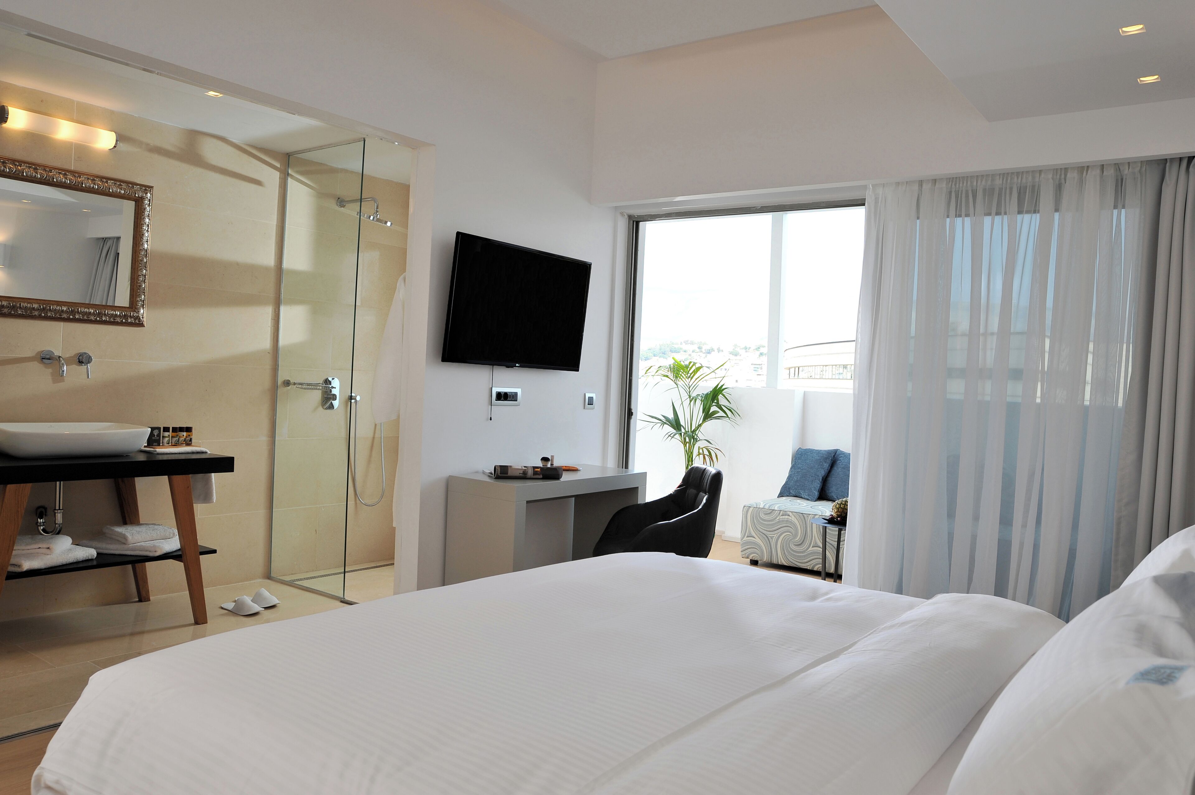A hotel room with a large bed, a desk, a television, a glass shower, and a balcony with curtains.
