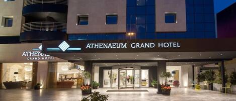 Entrada del Hotel Athenaeum Grand con puerta de vidrio, plantas en macetas y un vestíbulo bien iluminado.