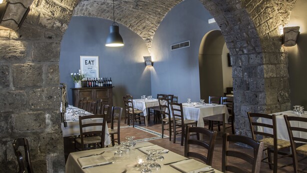 Un restaurante con arcos de piedra, mesas dispuestas con manteles blancos y botellas de vino en estanterías.