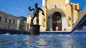 Een standbeeld in een fontein met waterstralen, voor een gebouw met de naam "KAUPPA-HALLI".