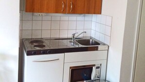 Een kleine keuken met houten kastjes, een spoelbak, een fornuis, een oven en een prullenbak.