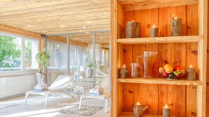 A spa area with wooden shelves holding candles, a pitcher of water, and a bowl of fruit. There are white lounge chairs and a large window with a view of greenery. - Arca Aparthotel & Spa (Zermatt)