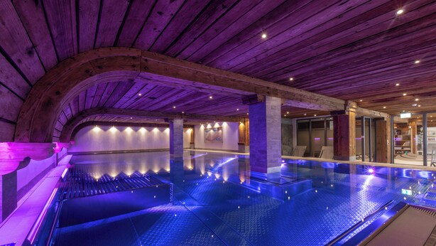A large indoor swimming pool with a wooden ceiling and walls, surrounded by glass doors and windows. - Arca Aparthotel & Spa (Zermatt)