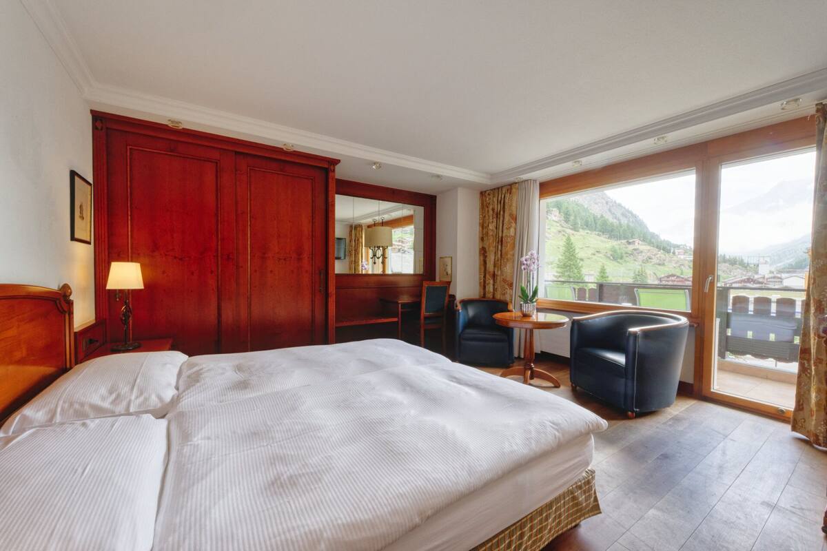 A hotel room with a large bed, a desk, a chair, and a view of a mountainous landscape.
