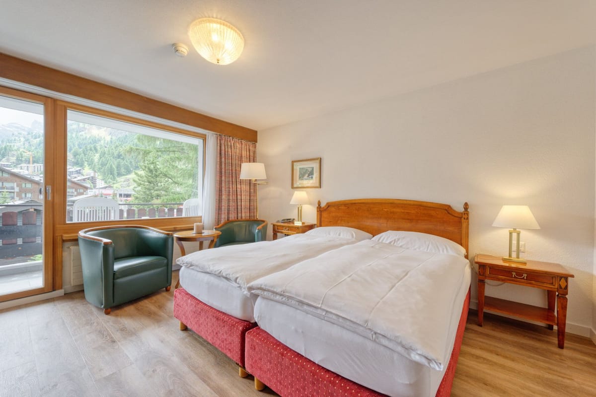 A hotel room with two beds, a window with a view of buildings and trees, a wooden headboard, and a small table with a lamp.