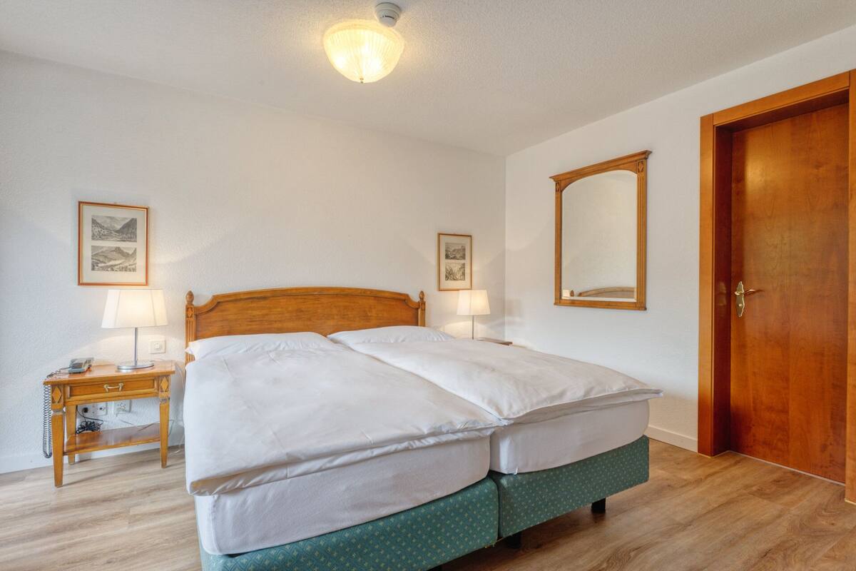 A double bed with a wooden headboard, two bedside tables with lamps, a wooden door, and two framed pictures on the walls.