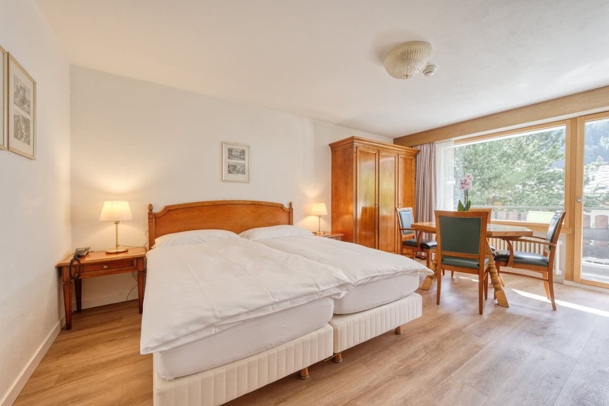 A hotel room with a double bed, bedside tables, a wooden wardrobe, a dining table with chairs, and a large window offering a view of the outdoors.