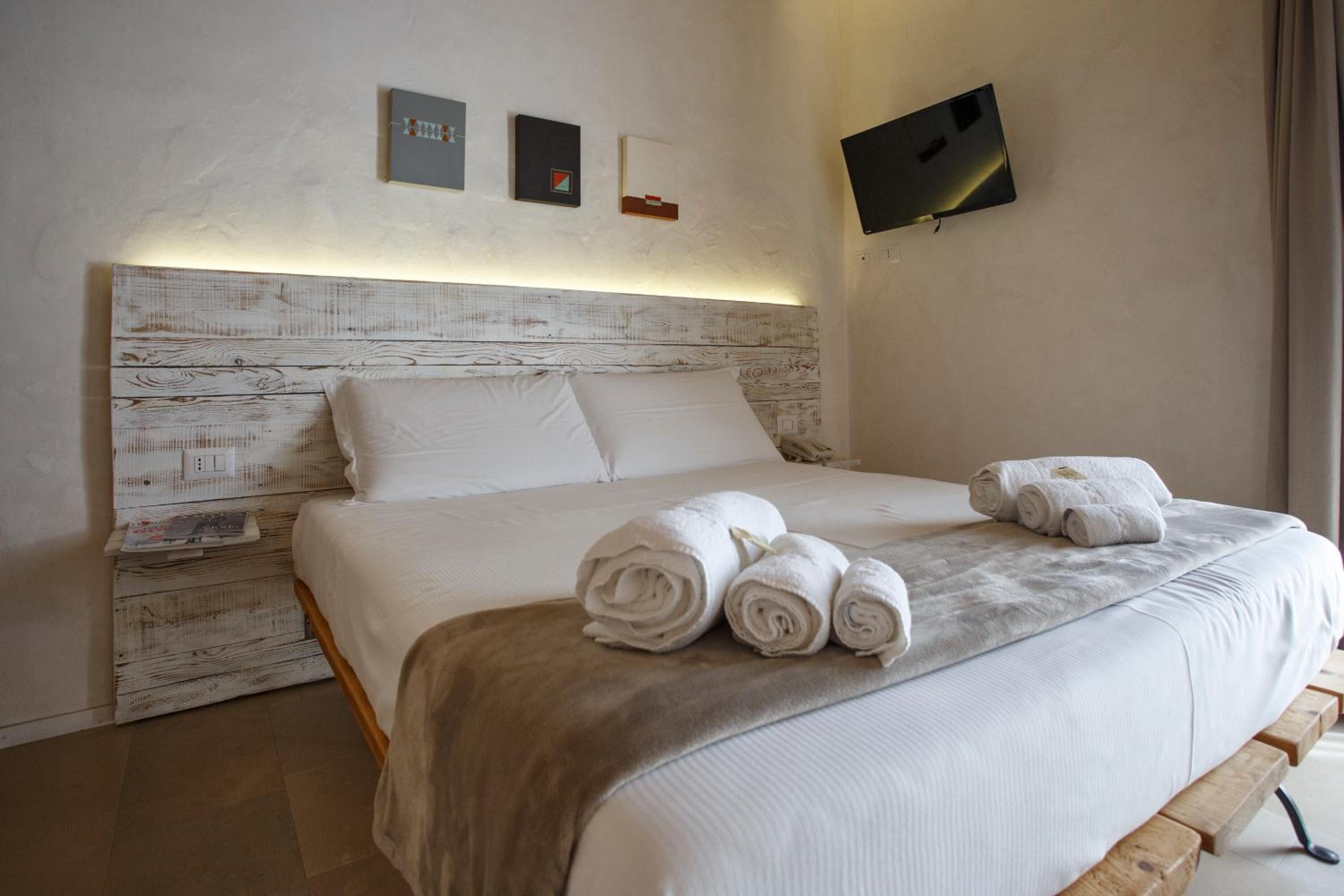 Una cama bien hecha con sábanas blancas y toallas enrolladas, un cabecero de madera y un televisor de pantalla plana montado en la pared.