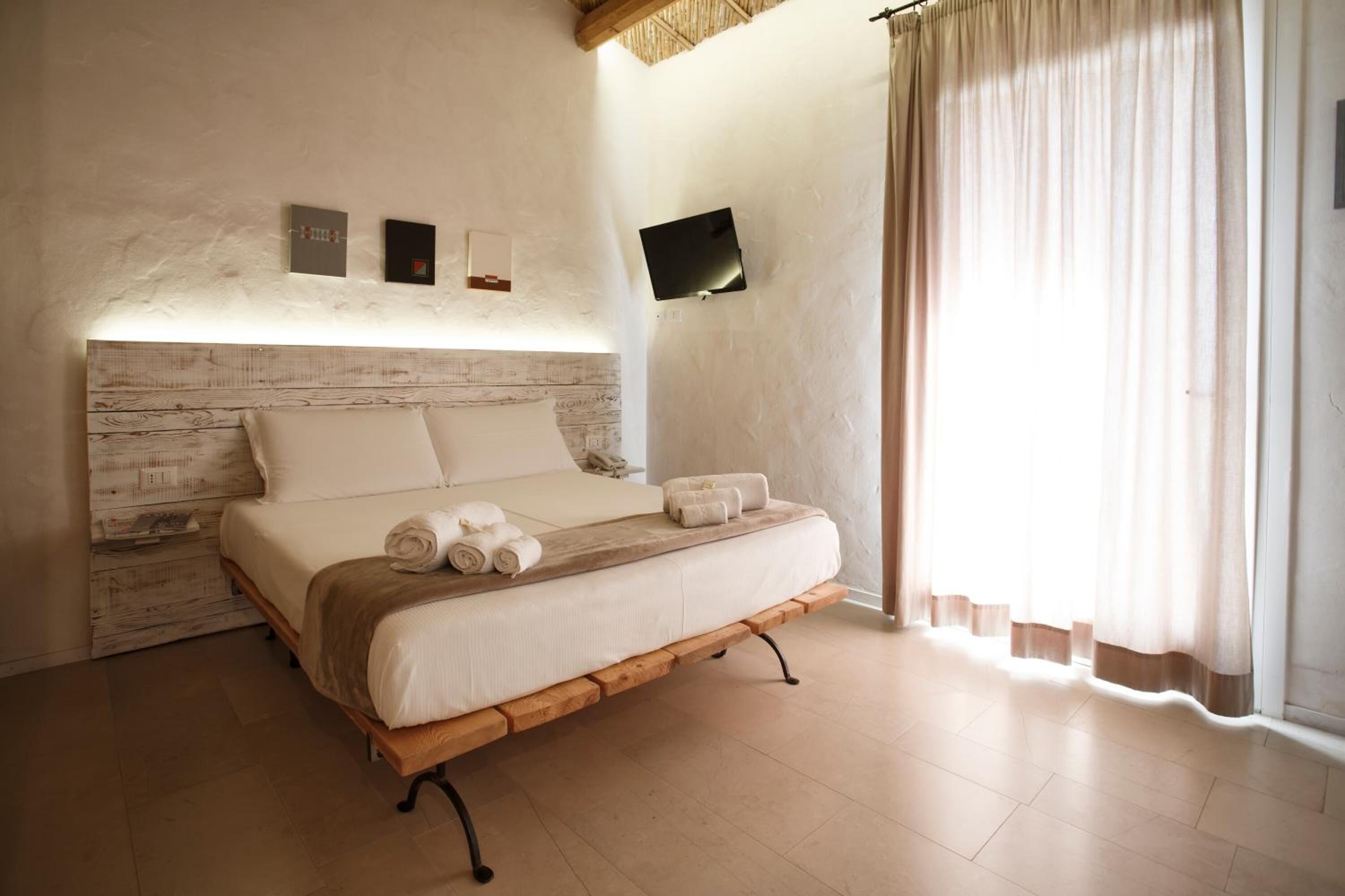 A bedroom with a wooden headboard, a bed with white linens, a flat-screen TV mounted on the wall, and a window with curtains.