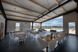 A spacious indoor dining area with multiple tables set for a meal, large windows offering a view of the outdoors, and a modern architectural design with exposed wooden beams and a high ceiling.