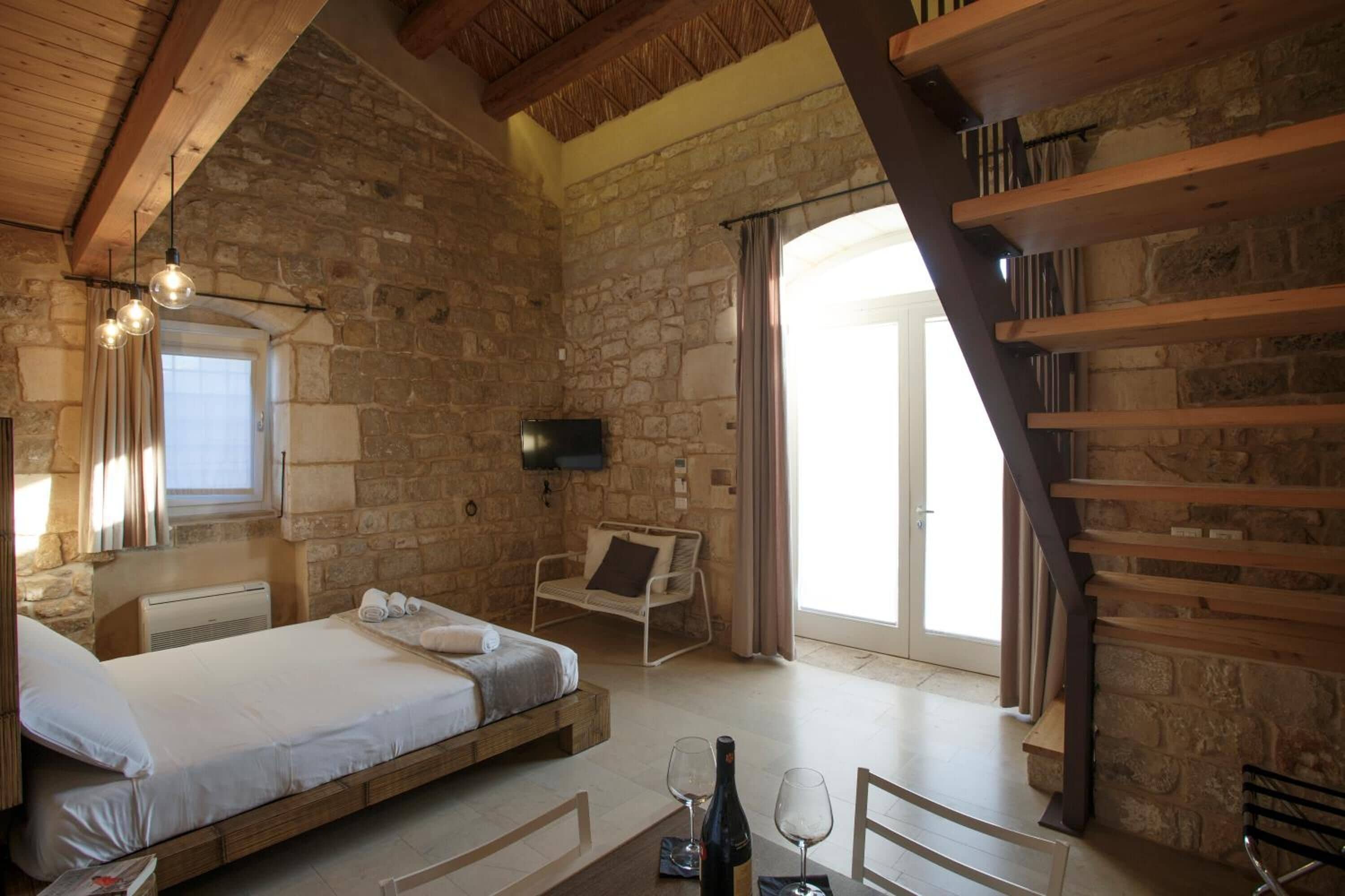 A rustic bedroom with a stone wall, wooden beams, a bed, a chair, and a table with a bottle and glasses.