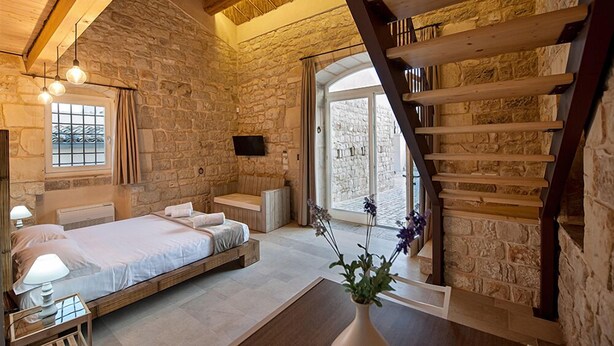 Une chambre avec un mur en pierre, un lit en bois, un banc, une grande fenêtre et un escalier.