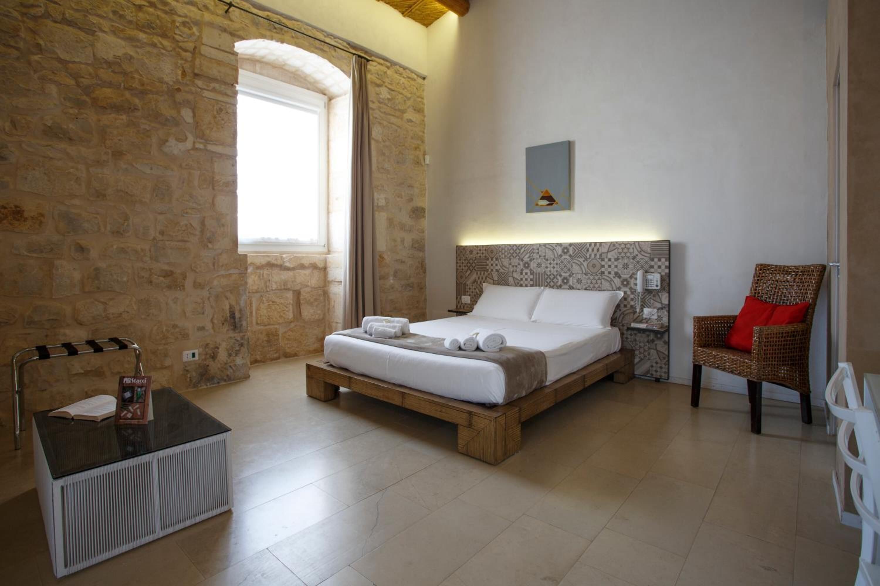 A bedroom with a stone wall, a bed with white linens, a wooden bedside table, a wicker chair with a red cushion, and a small table with a book and a menu.
