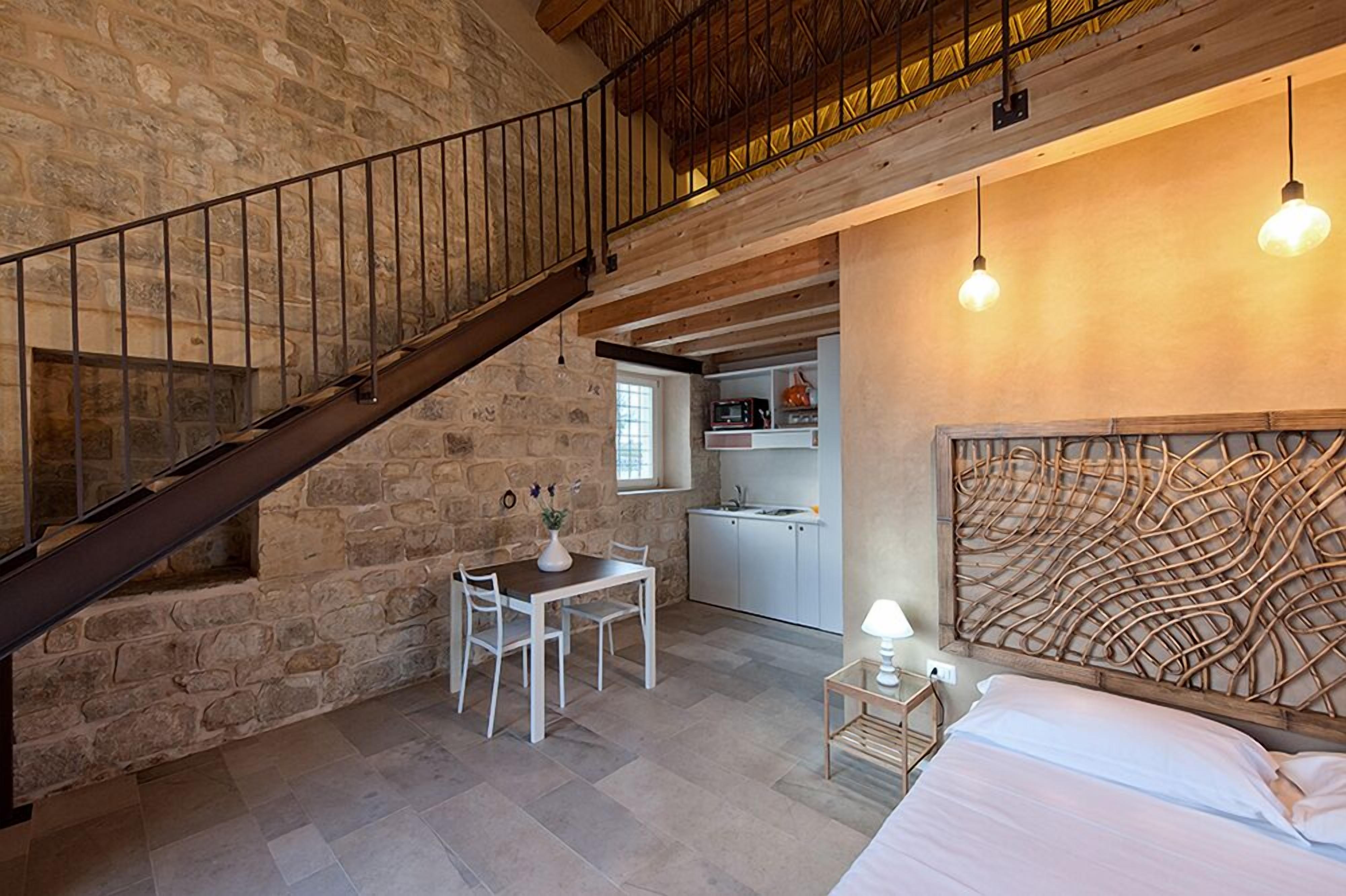 A rustic room with a stone wall, a wooden staircase, a dining area with a table and chairs, a kitchen, and a bed with a unique headboard.