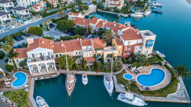 An aerial view of a marina with multiple boats docked, surrounded by residential buildings with red-tiled roofs and swimming pools. - Port Alacati Hotel (Cesme)