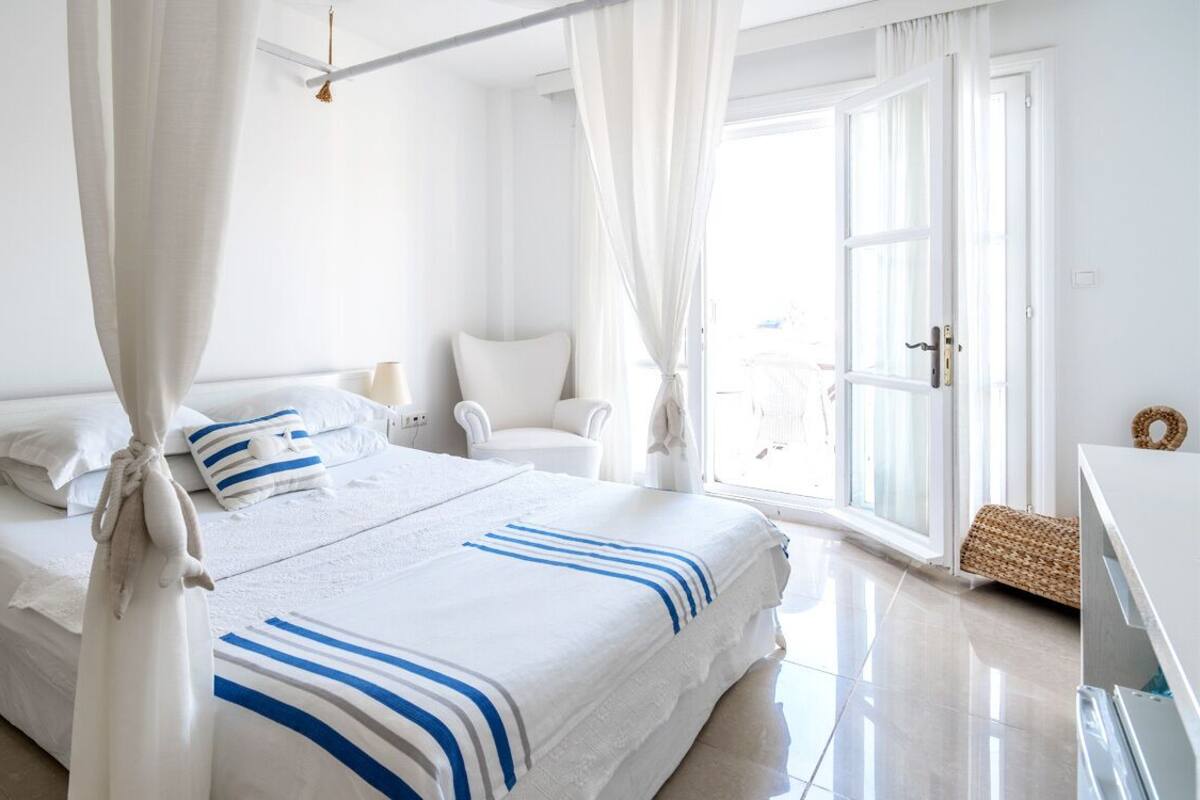 A hotel room with a large bed, white curtains, a white armchair, and a wicker basket.
