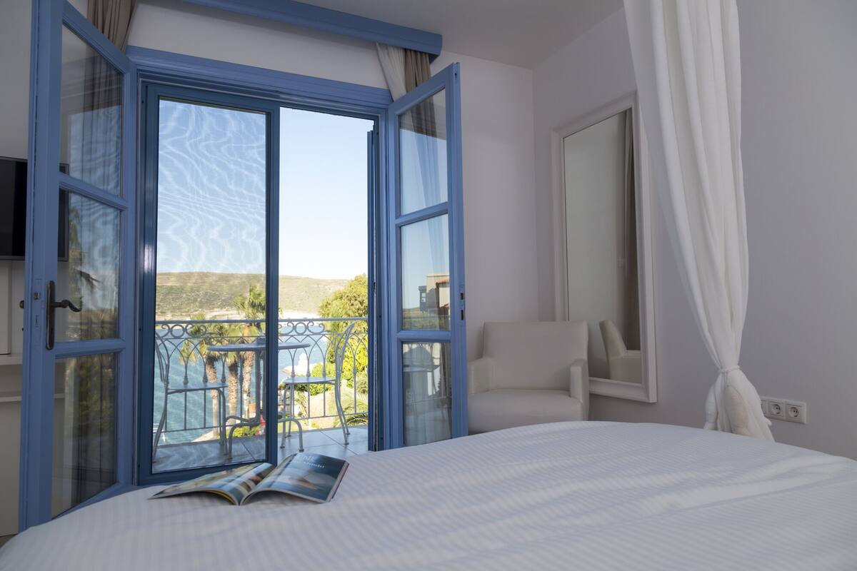 A hotel room with a large bed, a view of the sea, and a balcony with a chair.