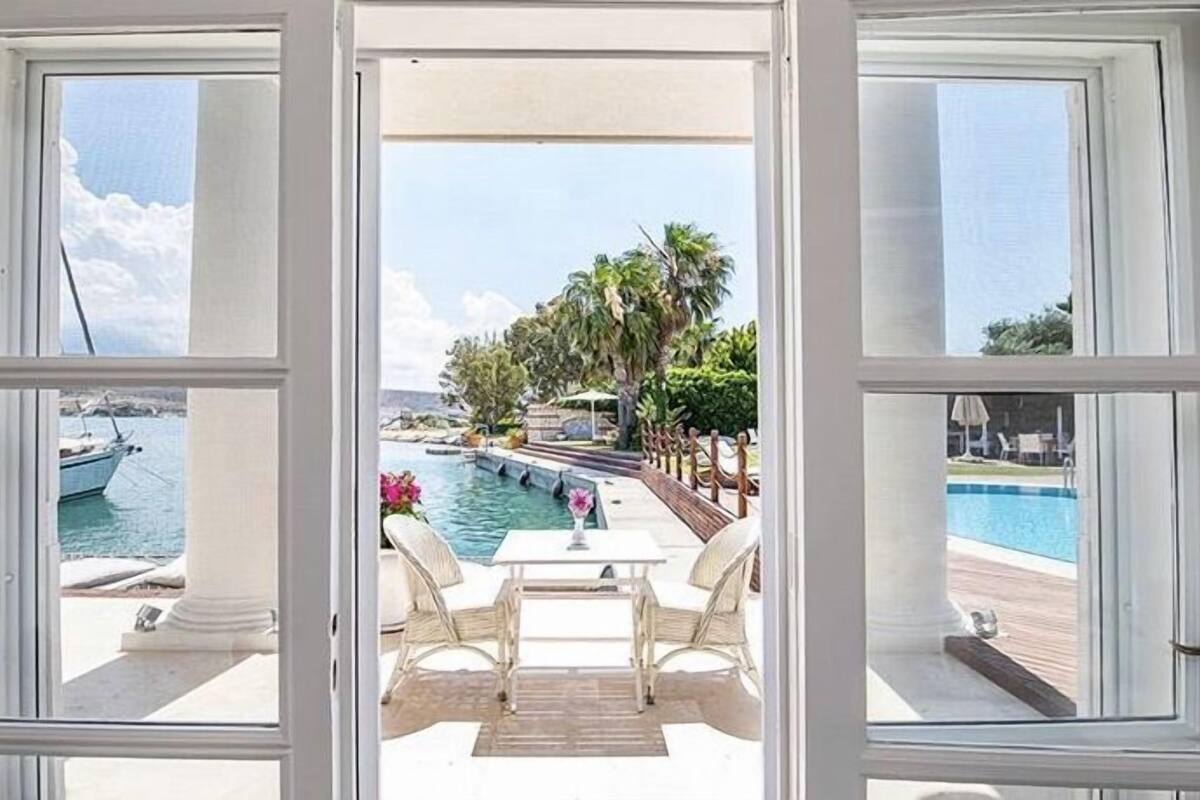 A view through a window of a pool area with a table and chairs, a pool, and a boat in the background.