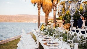 A long dining table set for a wedding with white chairs, glassware, and floral centerpieces. - Port Alacati Hotel (Cesme)