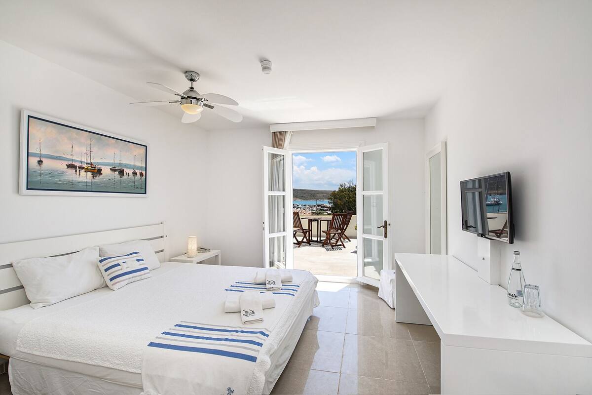 A hotel room with a bed, a desk, a TV, and a balcony with a view of boats and the sea.