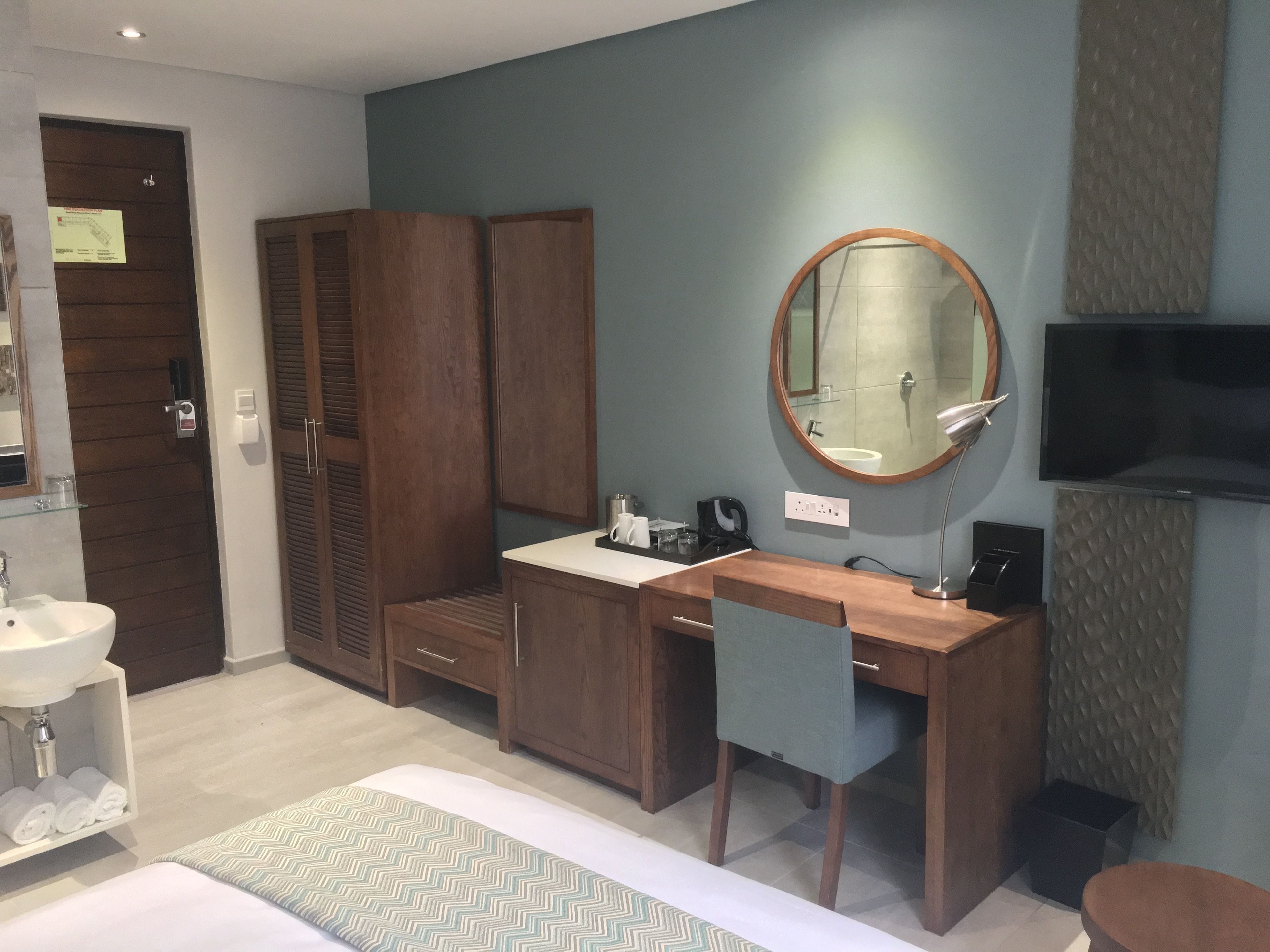 Una habitación de hotel con un escritorio de madera, un espejo redondo, un televisor montado en la pared, una cama con edredón verde y blanco, y un armario de madera.