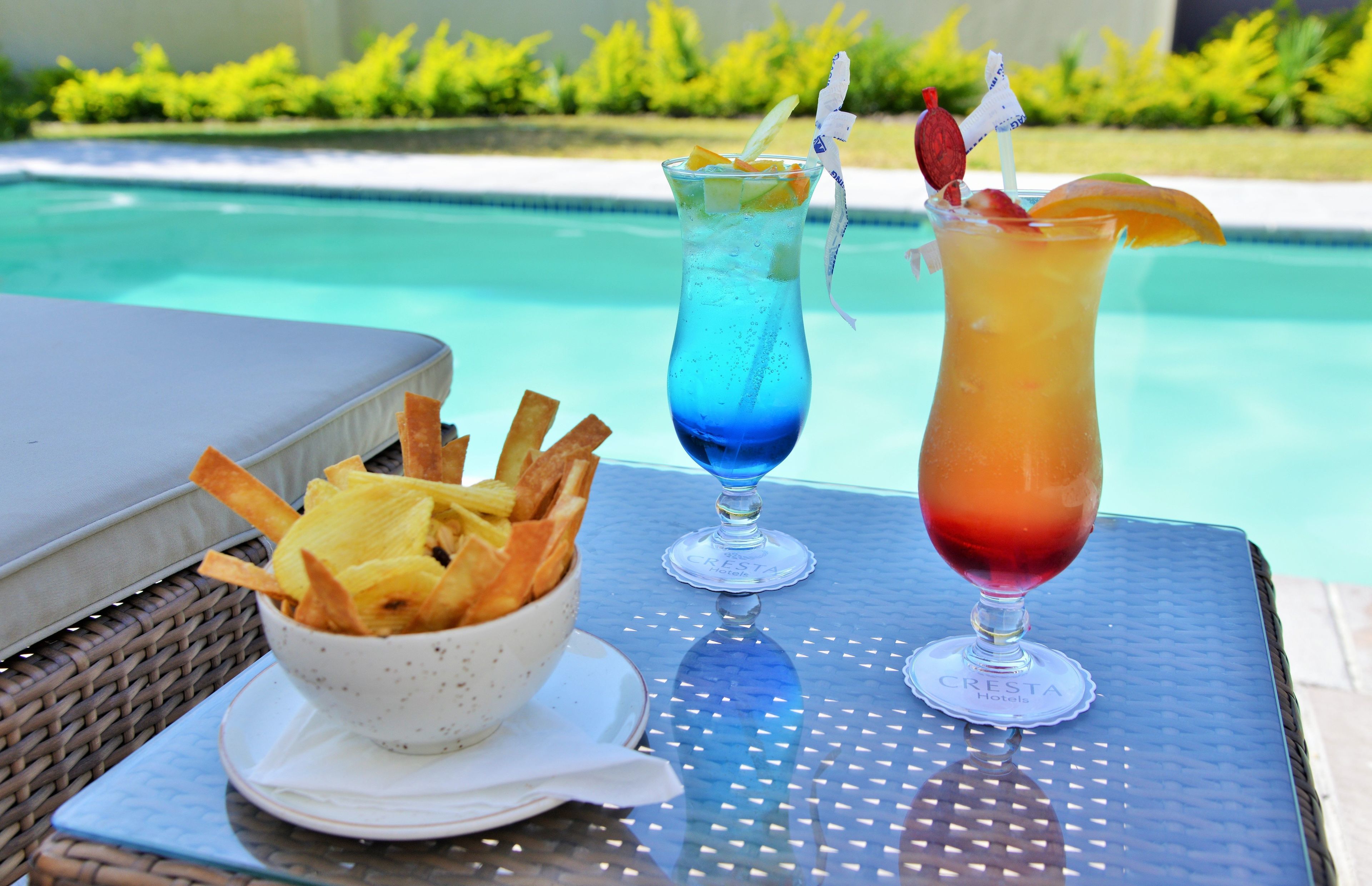 A poolside table with two drinks and a bowl of chips.