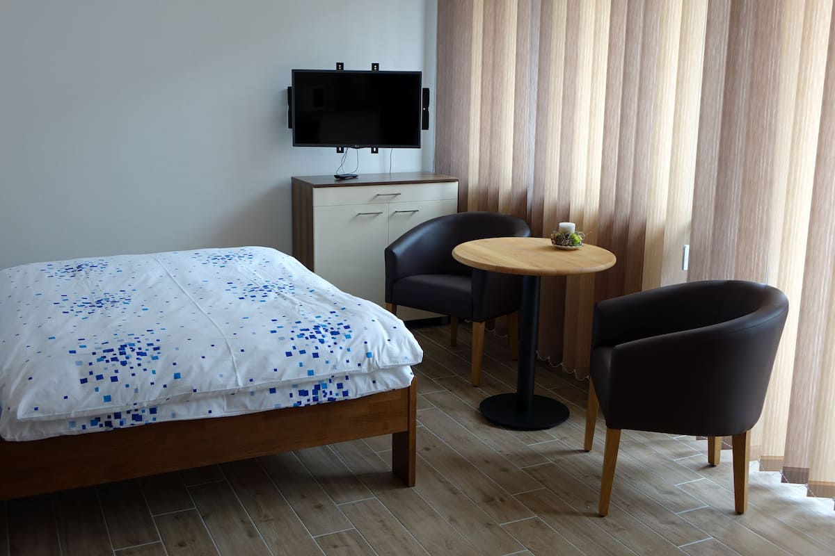 Habitación de hotel con cama, televisión montada en la pared, una mesita redonda con una silla y una ventana con cortinas.