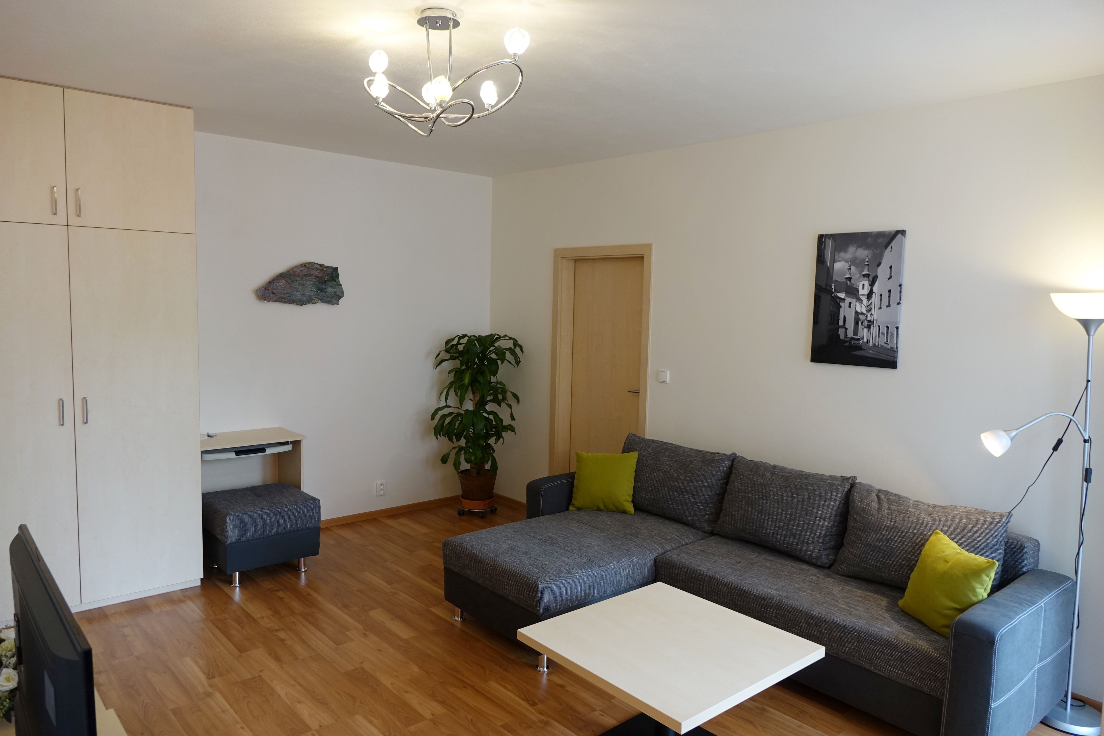 A modern living room with a grey sectional sofa, a white coffee table, a small blue armchair, a potted plant, and a black and white cityscape painting on the wall.