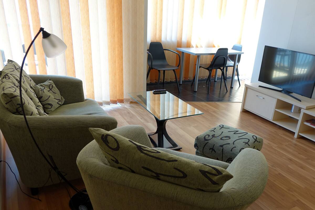 Una sala de estar con sofá, reposapiés, televisor y una zona de comedor con sillas y una mesa.