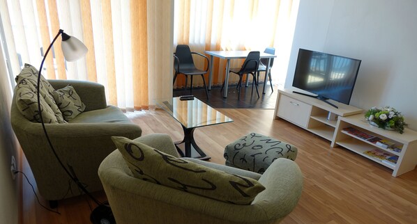 Uma sala de estar com sofá, um banquinho, televisão e um espaço de jantar com cadeiras e mesa.