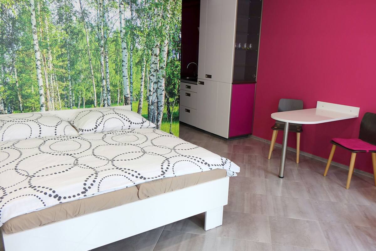 Un dormitorio con cama, una mesita, sillas, una kitchenette y vistas a árboles desde la ventana.