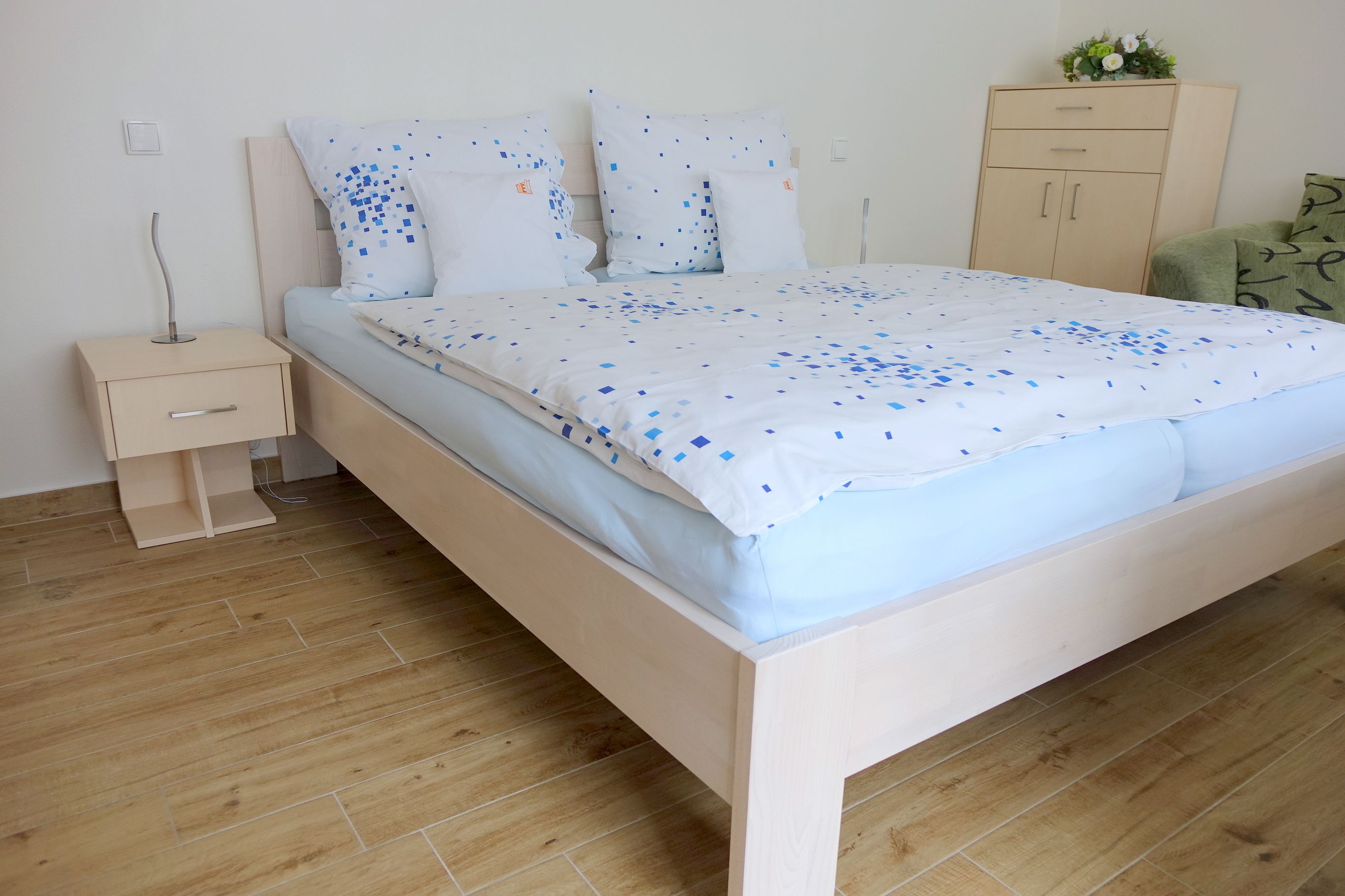 A neatly made bed with a patterned bedspread, a bedside table with a lamp, and a wooden wardrobe in a clean, minimalistic room.
